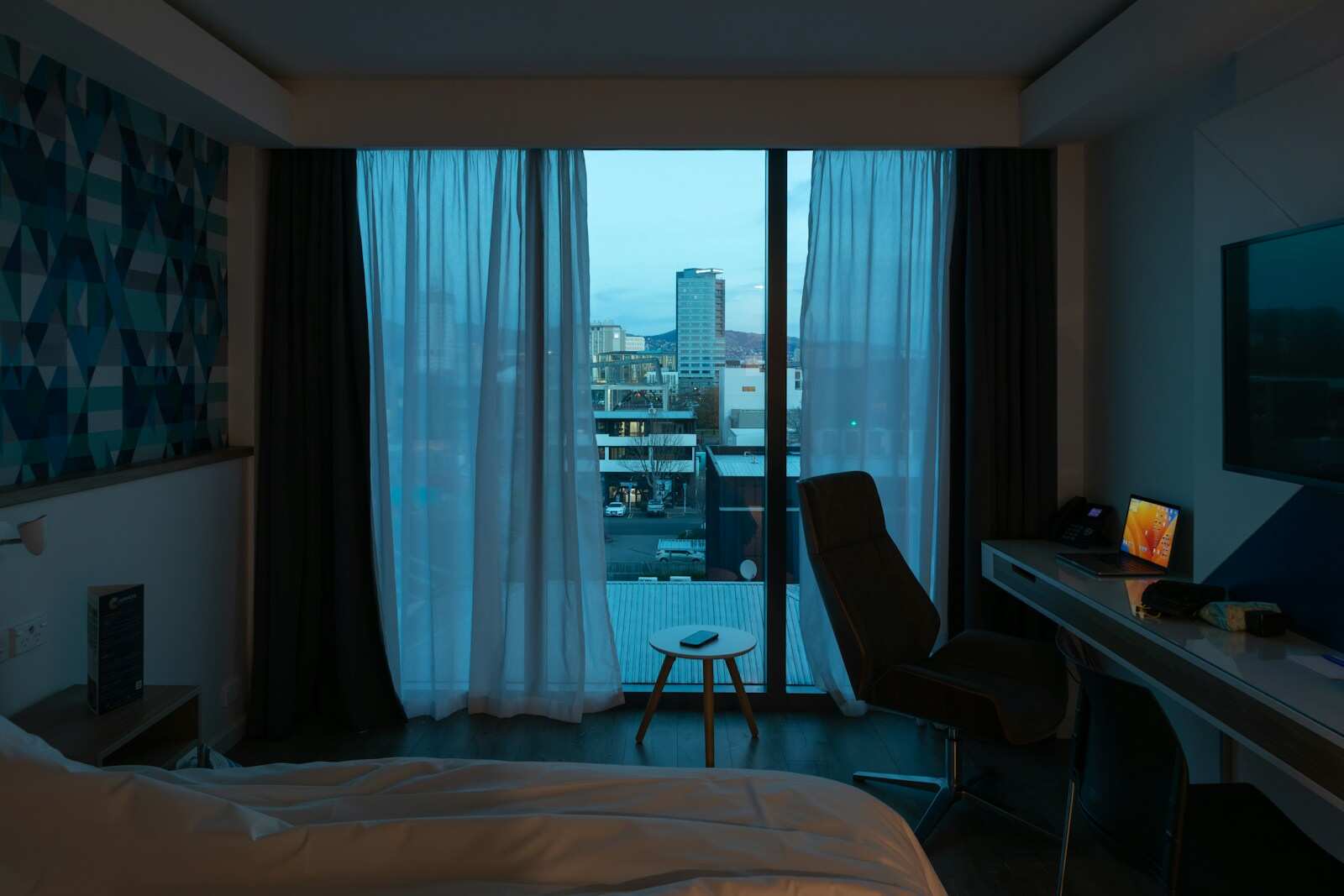 A bedroom at blue hour, the curtains open on a balcony and the city